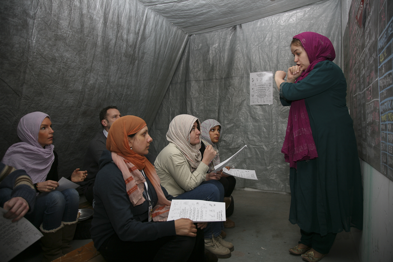 Women review documentation inside a temporary structure at a refugee camp, highlighting stories of migration.